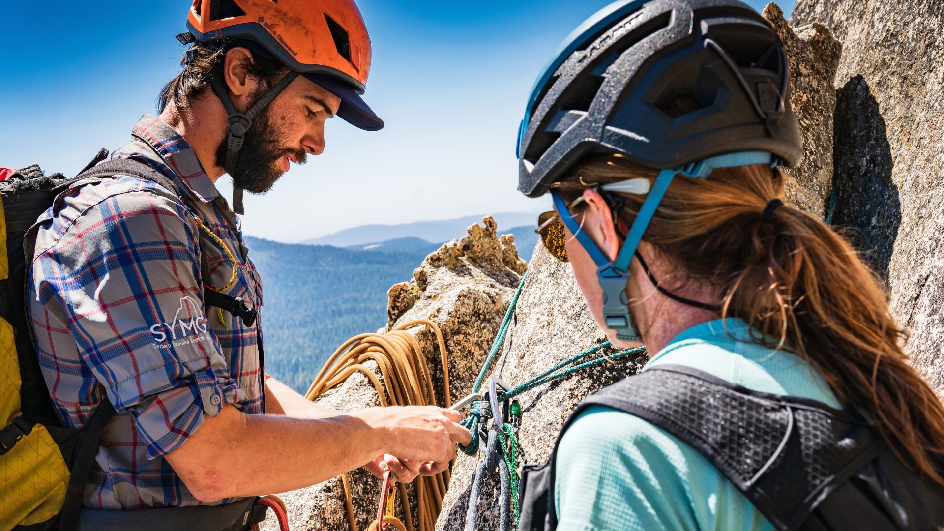 A climbing guide works on an anchor while another climber looks on