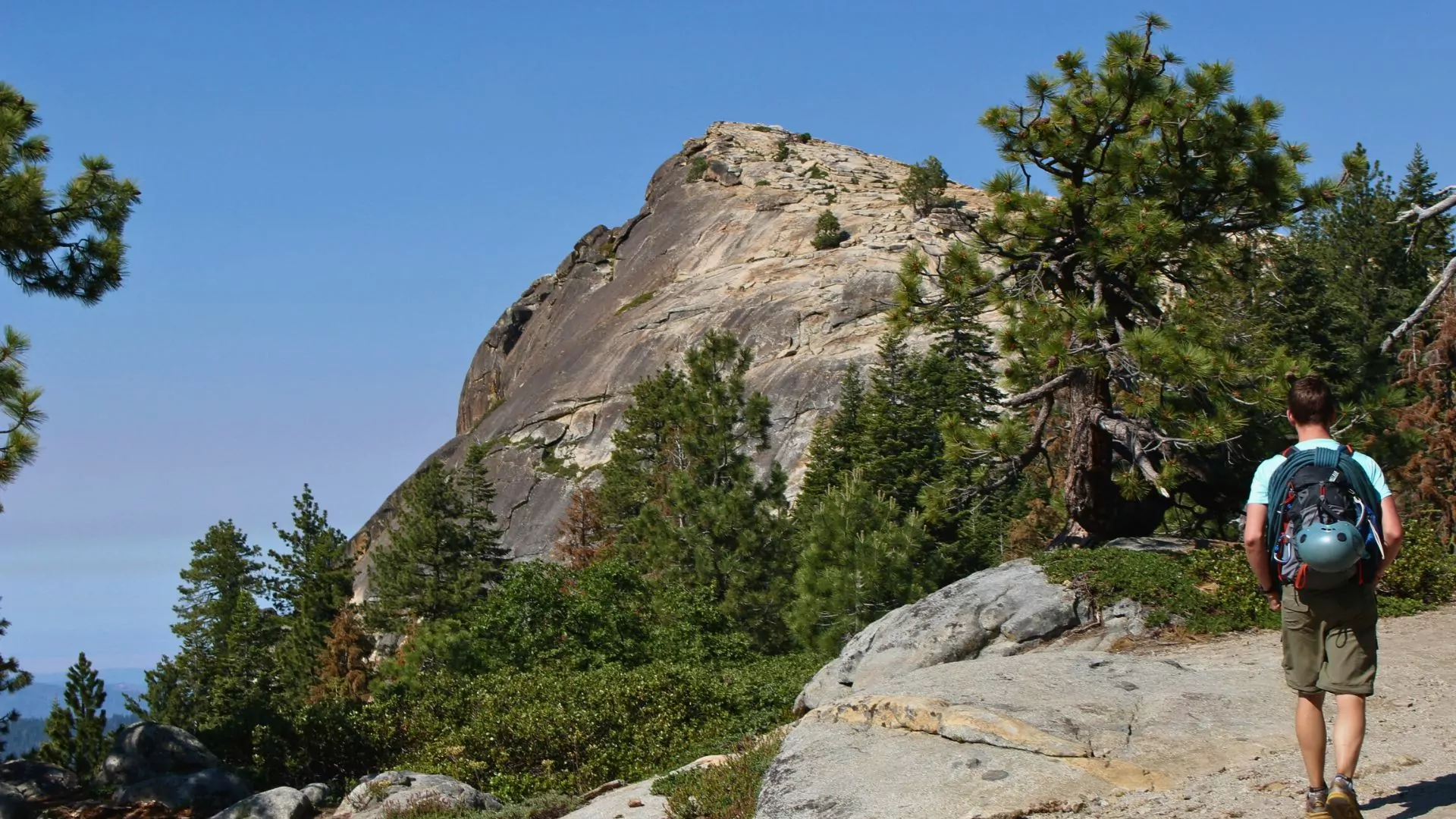 A man with a rope and helmet slung across his pack hikes toward a rocky mound