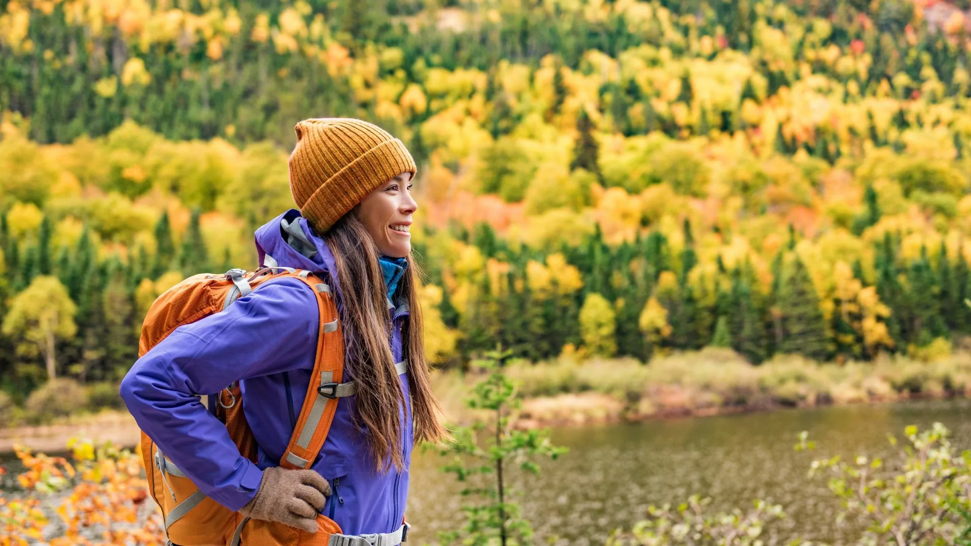 A woman wearing a daypack stands in front of a forest in full fall foliage