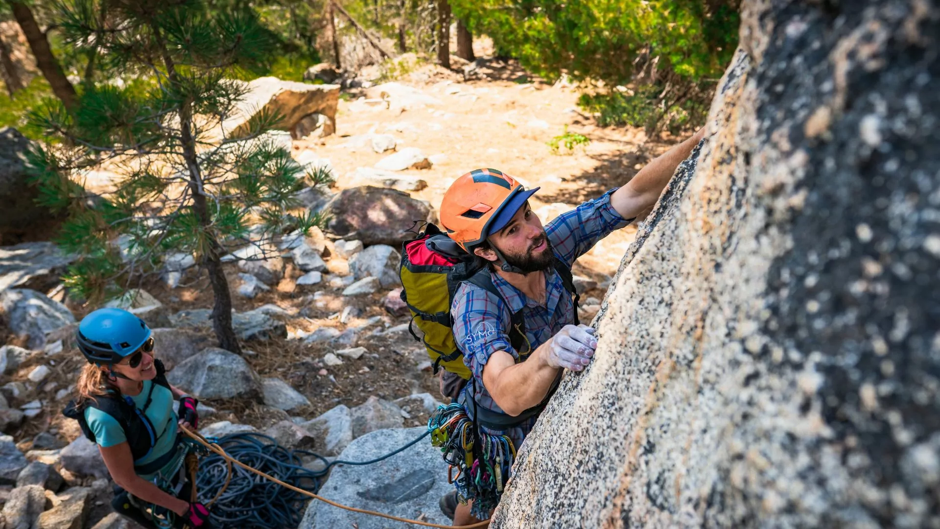 A man with a helmet and backpack lead climbs a granite face while a women belays him below