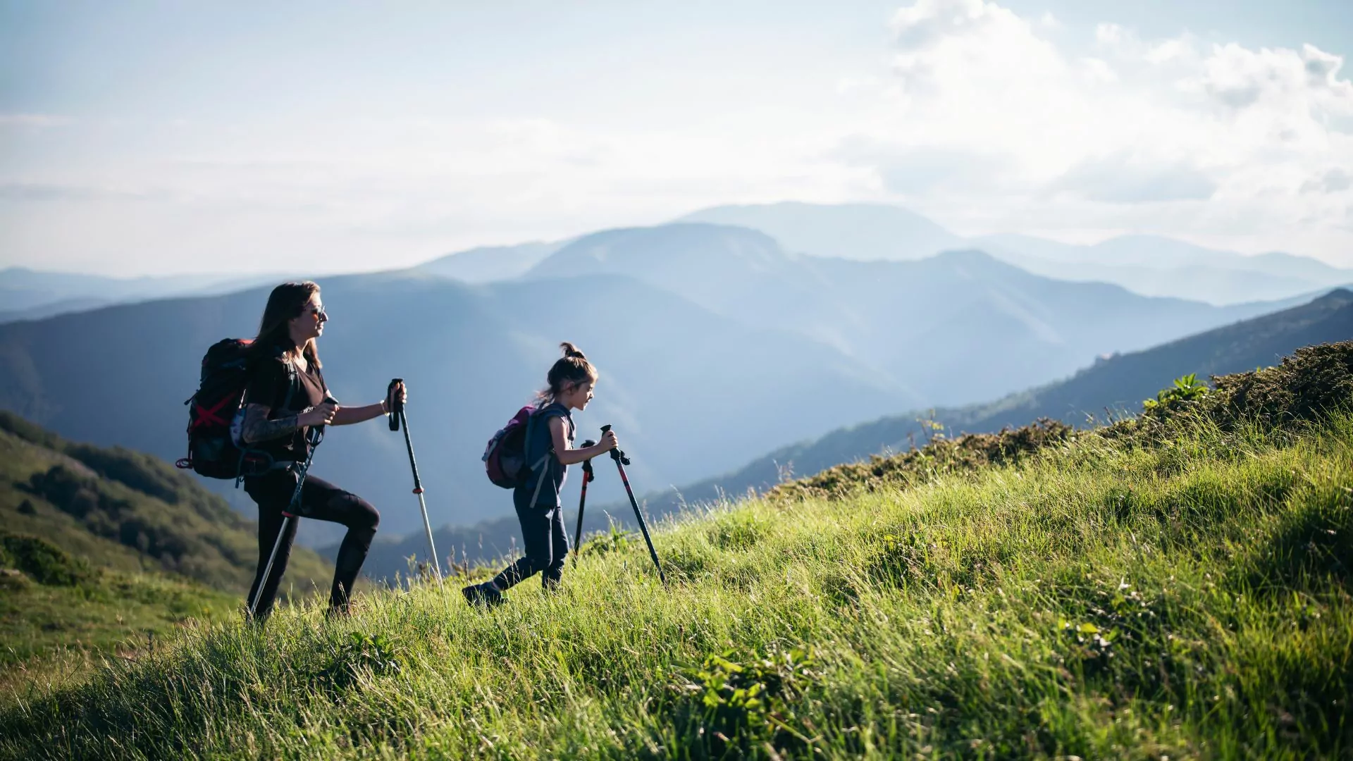 A woman and girl with daypacks and trekking poles hike through a field