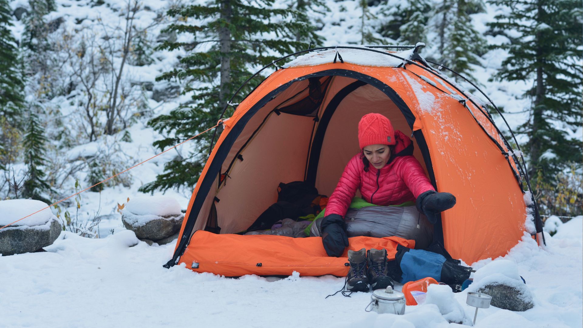 A woman in a coat and hat opens her tent door in the snow