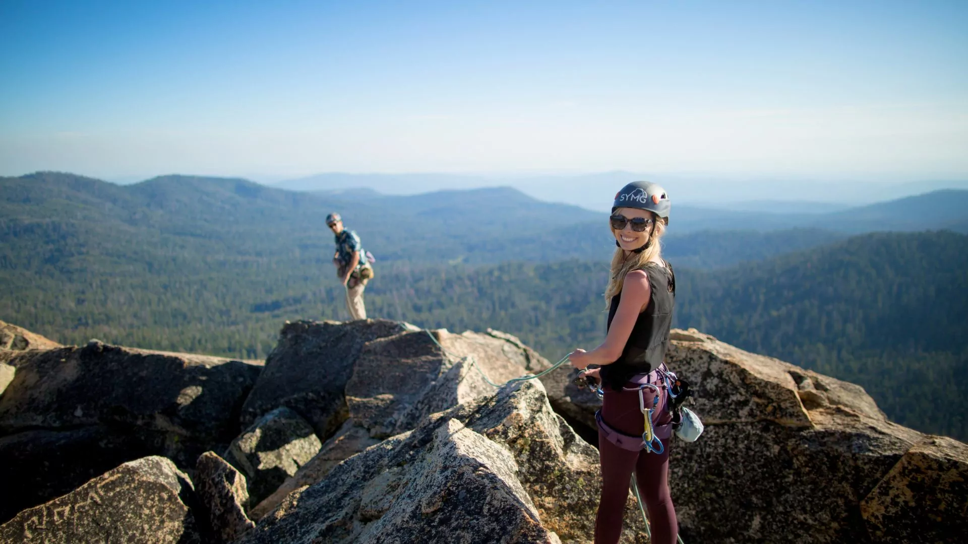 A woman with a helmet and harness on feeds rope to another climbing on a rocky summit